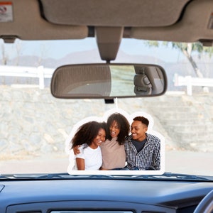 May include: A car's rearview mirror with a photo of three people smiling and hugging. The photo is attached to the mirror with a string.
