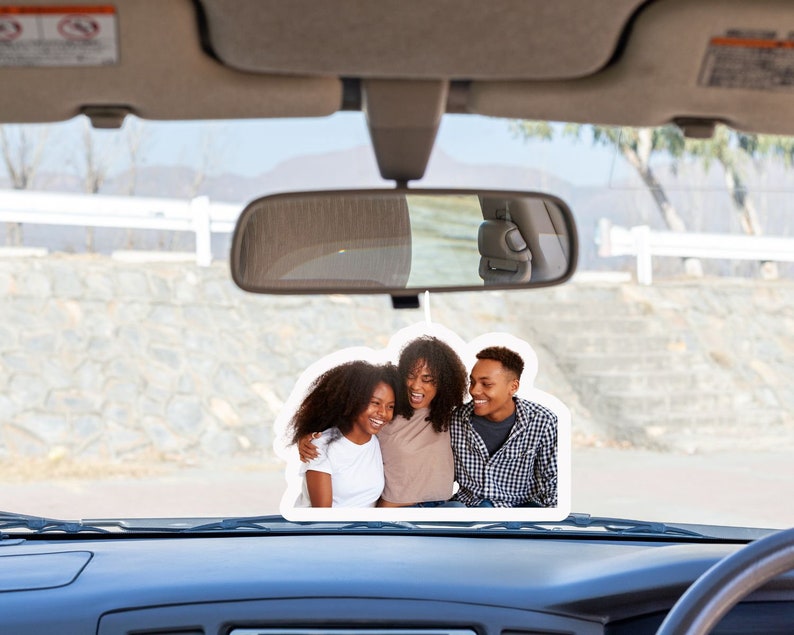 May include: A car's rearview mirror with a photo of three people smiling and hugging. The photo is attached to the mirror with a string.