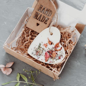 Puede incluir: Un adorno de cer&aacute;mica blanco en forma de coraz&oacute;n con flores y hierbas secas en su interior, empaquetado en una caja marr&oacute;n con una etiqueta "Hand Made".