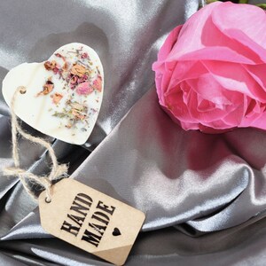 Puede incluir: Un derretido de cera blanco en forma de coraz&oacute;n con flores y hierbas secas, una rosa rosa y una etiqueta marr&oacute;n que dice "HAND MADE" sobre una tela de sat&eacute;n plateada.