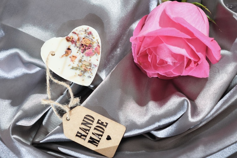 Puede incluir: Un derretido de cera blanco en forma de coraz&oacute;n con flores y hierbas secas, una rosa rosa y una etiqueta marr&oacute;n que dice "HAND MADE" sobre una tela de sat&eacute;n plateada.