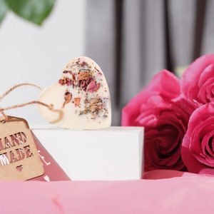Puede incluir: Una pastilla de jab&oacute;n blanca en forma de coraz&oacute;n con flores y hierbas secas incrustadas. El jab&oacute;n est&aacute; sobre una caja blanca con una etiqueta marr&oacute;n que dice "HAND MADE" con un peque&ntilde;o s&iacute;mbolo de coraz&oacute;n. El jab&oacute;n est&aacute; al lado de un ramo de rosas rosas.