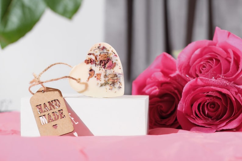 Puede incluir: Una pastilla de jab&oacute;n blanca en forma de coraz&oacute;n con flores y hierbas secas incrustadas. El jab&oacute;n est&aacute; sobre una caja blanca con una etiqueta marr&oacute;n que dice "HAND MADE" con un peque&ntilde;o s&iacute;mbolo de coraz&oacute;n. El jab&oacute;n est&aacute; al lado de un ramo de rosas rosas.