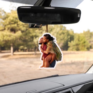 May include: A car air freshener featuring a photo of a couple embracing. The woman is wearing a floral patterned shirt and the man is wearing a red shirt. The photo is set against a blurred background of trees and a dirt road.