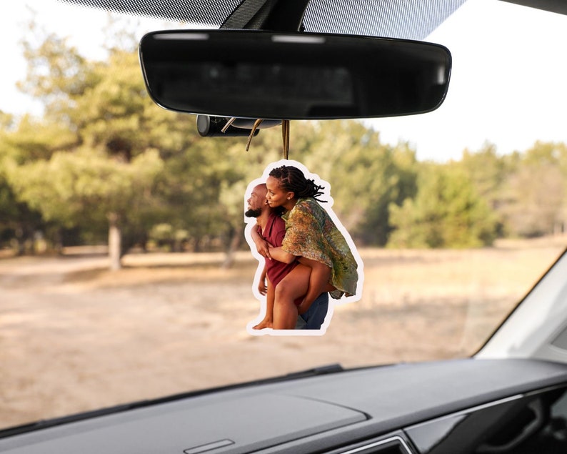 May include: A car air freshener featuring a photo of a couple embracing. The woman is wearing a floral patterned shirt and the man is wearing a red shirt. The photo is set against a blurred background of trees and a dirt road.