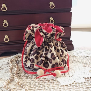 May include: A small drawstring bag with a leopard print pattern, red lining, and a red drawstring. The bag is made of fabric and has two wooden beads on the drawstring. A pearl necklace is in the foreground.