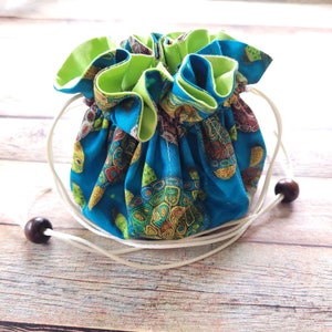 May include: A blue drawstring bag with a green lining and a turtle print. The bag has a white drawstring and brown wooden beads.