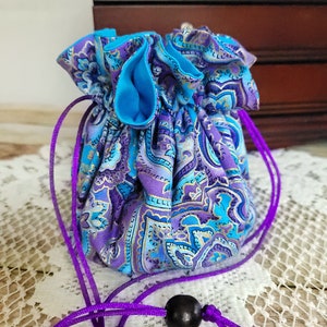 May include: A small, drawstring pouch with a paisley pattern in shades of purple, blue, and gold. The interior is a solid turquoise blue. The drawstring is a vibrant purple, and a dark wooden bead is attached.
