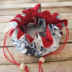 May include: A round, floral print drawstring bag with a red ruffled trim. The bag features a light gray background with dark gray and white floral patterns. Red drawstrings with wooden beads are visible.