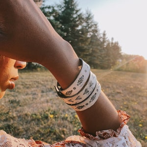 May include: Three white leather cuff bracelets with black etched designs are stacked on an arm. The bracelets feature floral and geometric patterns. The person is wearing a patterned dress with a lace sleeve. The background is a field with trees.