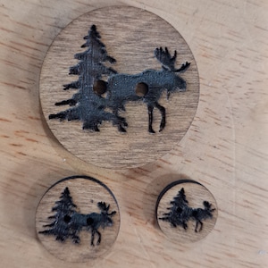 May include: Three wooden buttons with a moose and pine tree design. The largest button is approximately 2 inches in diameter and the smaller buttons are approximately 1 inch in diameter.