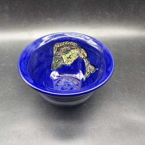 May include: A blue ceramic bowl featuring a hand-painted fish design. The fish is brown, green, and gold with a black eye. The bowl is ideal for serving snacks or desserts.