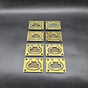 May include: Eight brass flush mount drawer pulls. Each pull has a rectangular base with a curved handle. The pulls are a warm gold colour and are arranged in two rows of four on a dark background.