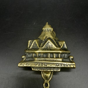 May include: A brass door knocker featuring a building design, with the words "OLD YARN MARKET DONCASTER" inscribed. The knocker has an aged appearance and a loop for attachment, reflecting a vintage aesthetic.