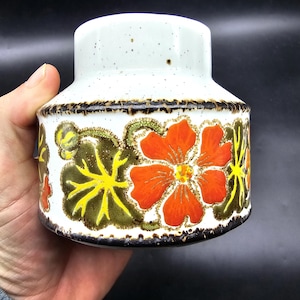 May include: A white ceramic jar with a speckled glaze and a floral design. The flowers are orange, yellow, and green. The jar has a brown rim.
