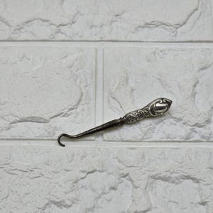 May include: A silver-toned button hook featuring an ornate, embossed handle with intricate scrollwork patterns and a smooth, tapered metal shaft that terminates in a small, curved hook designed for fastening buttons on clothing items.