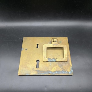 May include: A rectangular brass drawer pull with a keyhole and a rectangular handle. The brass has a patina with some blue-green oxidation. The handle is hinged and has a rectangular opening. The keyhole is oval-shaped.