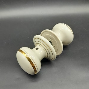 May include: Three ivory door knobs with gold bands. The knobs are oval and have a classic design. The hardware is likely for a door or cabinet. The measurements are in centimetres.