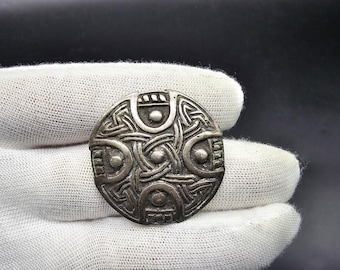 Pode incluir: Um broche circular de prata com um design de nó celta e pontos em relevo. O broche tem aproximadamente 5 cm de diâmetro.