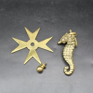 May include: A selection of brass decorative hardware components. Includes a Maltese cross, a seahorse, and a small knob with a threaded post. The items are displayed on a dark surface.