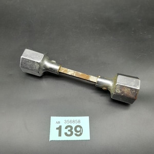 May include: A silver metal tool with hexagonal ends and a central, rectangular, rusty-coloured shaft. The tool is lying on a dark surface. A small, blue tag with the numbers "139" is visible.
