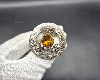 May include: A silver-toned brooch with a central amber-colored stone. The brooch features a circular design with detailed engravings and a thistle motif, showcasing a classic and elegant style.