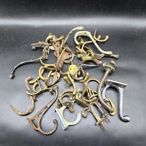 May include: A selection of antique metal hooks in diverse designs and finishes, such as brass, bronze, and silver. The hooks vary in shape and size, some with screws, ideal for hanging items.
