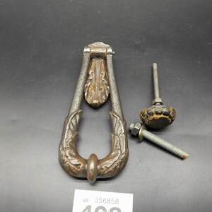 May include: A silver metal door knocker with a leaf design. The knocker is in the shape of a teardrop and has a round ring at the bottom. The knocker is attached to a wooden door.