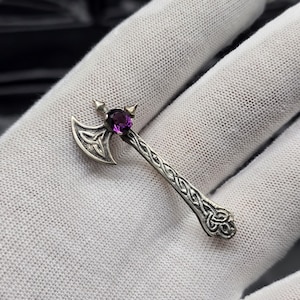 Puede incluir: Un broche de metal plateado con forma de hacha de batalla, que presenta un intrincado patrón de nudos celtas en el mango y la hoja. Una sola piedra preciosa morada, redonda y facetada, está engastada en la unión de la cabeza y el mango.