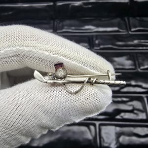 Könnte beinhalten: Silberfarbene Brosche mit einem Golfschläger, einer Distel mit einem lila Edelstein und einem Golfball. Die Brosche wird von einer behandschuhten Hand vor einem dunklen, strukturierten Hintergrund gehalten. Das Design ist detailliert.