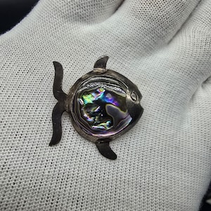 Puede incluir: Un broche de plata en forma de pez con una incrustación de nácar de abulón iridiscente. El pez tiene un diseño detallado y un pequeño ojo.