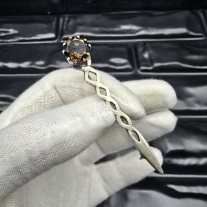 May include: A silver-toned hair pin with a large, faceted, brown gemstone at the top. The pin has a decorative, openwork design and a pointed end. The gemstone is round and has a dark, smoky appearance.