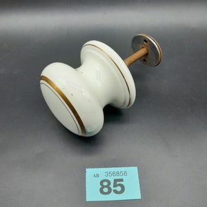 May include: White ceramic door knob with gold trim and a metal mounting plate. The knob is attached to a metal screw. The image also includes a blue price tag with the text "AB 356858 85".