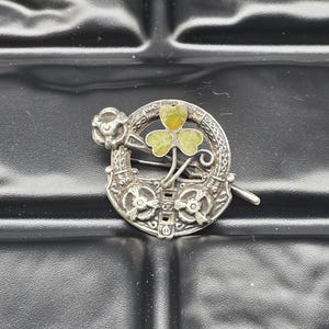 Può includere: Spilla argentata con design circolare e un trifoglio a tre foglie verdi al centro. La spilla ha un bordo decorativo e una piccola rosa sul lato sinistro. Design tradizionale.