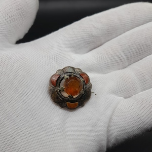 May include: A silver brooch featuring a round, orange gemstone set within a silver frame, accented by three smaller red and orange gemstones. The brooch has an intricate design.