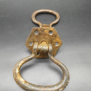 May include: An aged door knocker, featuring a hexagonal backplate and two rounded handles. The metal has a rusty, textured appearance, indicating age and use. The knocker is hinged, allowing the handle to swing and strike the door. The overall design is vintage.