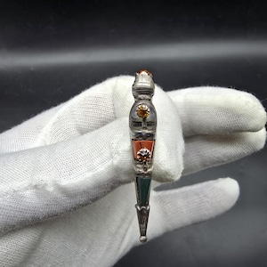 May include: A decorative silver dirk with a detailed design. The dirk features a brown gemstone at the top, with sections of orange, green, and silver. The handle has intricate patterns and a small, round tip.