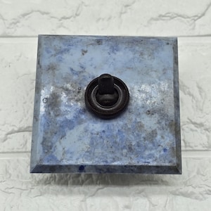 May include: A square light switch plate featuring a mottled blue and grey stone-like finish with a centred dark brown circular toggle switch. The plate has bevelled edges and a smooth, polished surface texture surrounding the central mechanism.