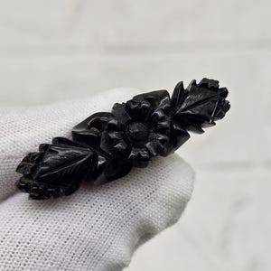 Può includere: Una spilla floreale nera con fiori e foglie scolpite. La spilla è realizzata in un materiale scuro e presenta un design dettagliato. La spilla è tenuta da una mano guantata su uno sfondo bianco.