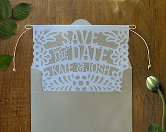 May include: A white paper save the date card with a cut-out design. The card says "Save the Date" with "Kate & Josh" underneath. The card is in a brown envelope.