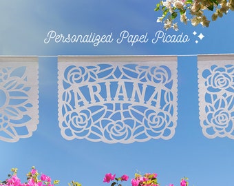 May include: White personalized papel picado banner with the name "Ariana" cut out. The banner is strung against a bright blue sky, with pink and white flowers in the foreground and background. The text "Personalized Papel Picado" is above the banner.