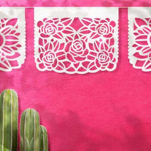 K&ouml;nnte beinhalten: Wei&szlig;e Papierschnitte mit floralen Designs h&auml;ngen vor einem leuchtend pinkfarbenen Hintergrund. Die Ausschnitte haben die Form eines Banners und zeigen komplizierte Blumenmuster.