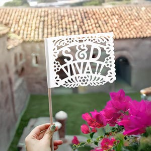 May include: White paper flag with a cut-out design featuring the text "S&D Viva!" in a decorative font. The flag is held by a hand against a backdrop of green foliage and a building with a terracotta roof.