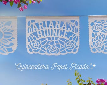 May include: White paper banners with cut-out designs, including roses and the text "Ariana's Quince". The banners are strung together against a blue sky with pink flowers in the foreground and background.
