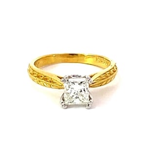 May include: A gold ring with a square, clear gemstone at the center. The band has an engraved leaf design. The ring is set against a white background.