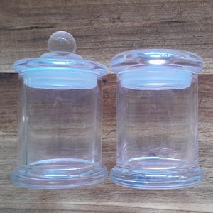 May include: Two clear glass jars with lids. The jars are cylindrical and have a smooth, rounded bottom. The lids are made of a clear, frosted glass.