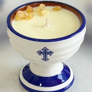 May include: A ceramic candle holder in white and blue, featuring a cross motif. The candle has a creamy yellow wax, a single wick, and amber crystals. The rim is brown, and the base is blue and white. A decorative item.
