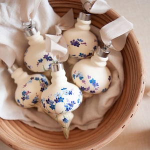 Peut inclure: Un ensemble de six boules de Noël en verre blanc peintes de fleurs bleues de myosotis. Les boules sont dans un panier en osier avec un tissu blanc.