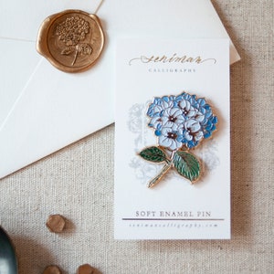 K&ouml;nnte beinhalten: Eine blaue und wei&szlig;e Emaille-Anstecknadel mit einer Hortensienbl&uuml;te und gr&uuml;nen Bl&auml;ttern. Die Anstecknadel befindet sich auf einer wei&szlig;en Karte mit dem aufgedruckten Text "Senimar Calligraphy" und "Soft Enamel Pin".