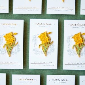 May include: A set of eight yellow daffodil enamel pins on white cards with the text "Seniman Calligraphy" and "Soft Enamel Pin" printed on them. The cards are arranged in a grid pattern on a green surface.